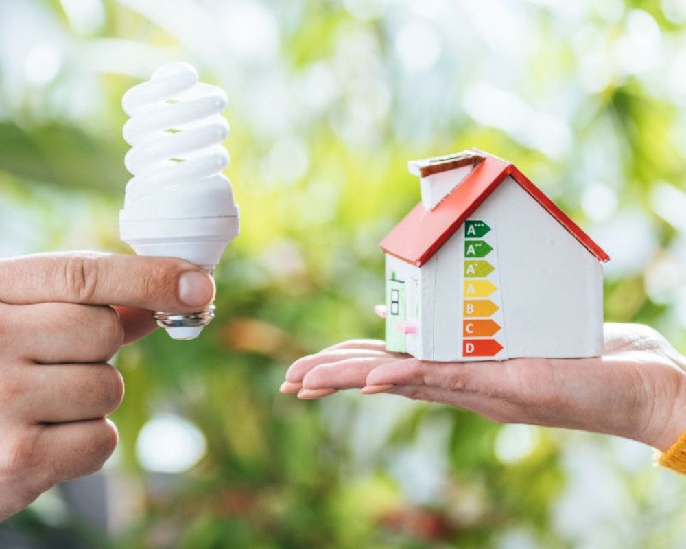 Imagen que representa ahorro en la factura de la luz. En una mano maqueta de una casa y en la otra una bombilla de led.